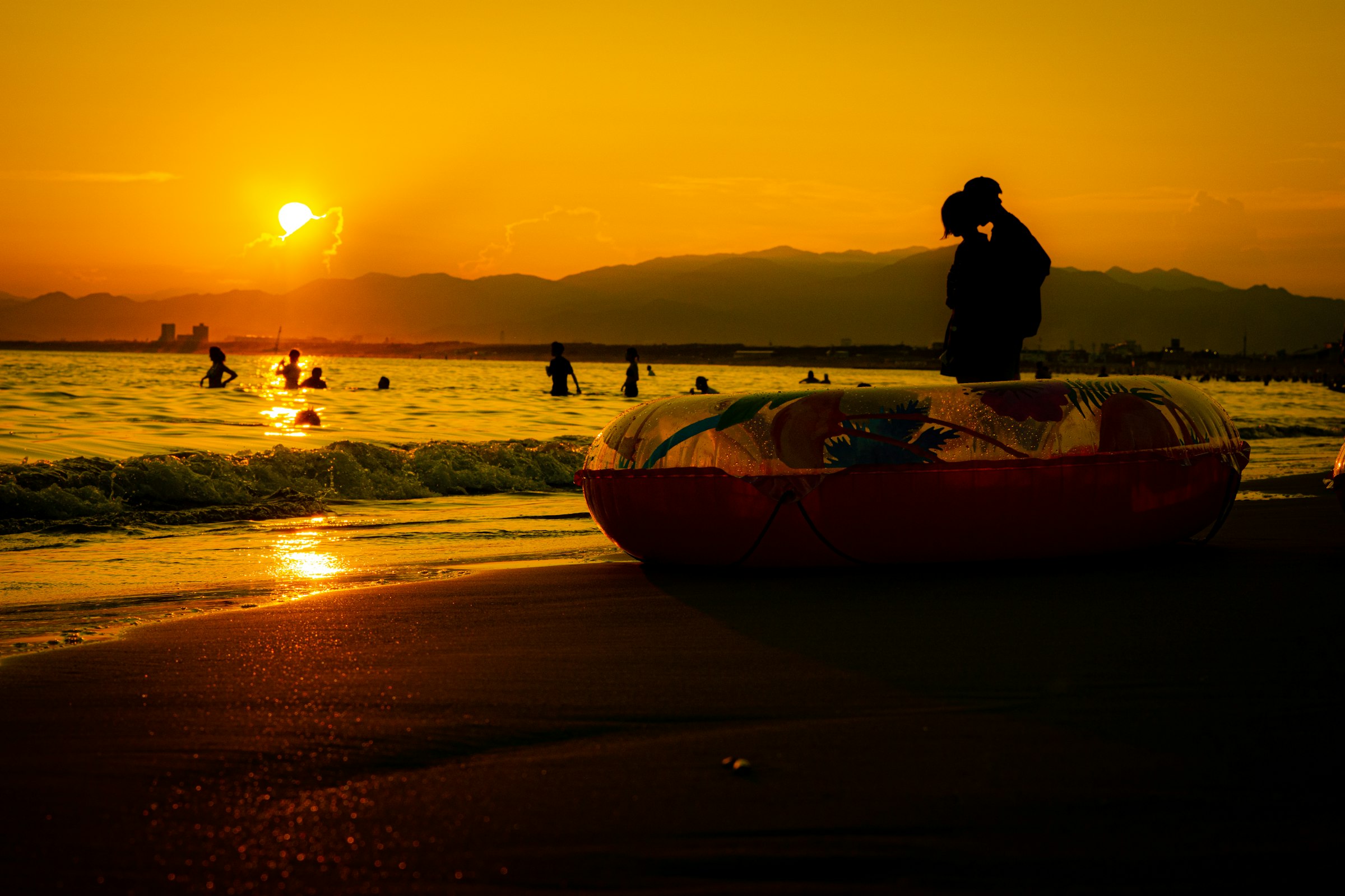 夕暮れの浜辺で、寄り添うように抱き合う二人のシルエット。奥には夕日と山並みが見える